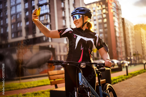 Cheerful woman taking selfie with yellow smartphone posing with bicycle. Mobile photography and modern social media communication concept.