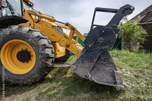 A loader is parked in a yard, ready to dig soil and lift materials. The machine sits near a house with green grass around it on a cloudy day