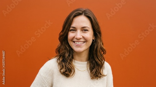 A woman with long brown hair, wearing a white sweater, standing in front of an orange background.