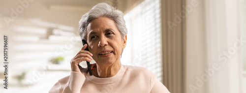 Senior woman uses smartphone for serious conversation at home. Indoor communication makes concerned attentive elderly lady thoughtful while listening to voice call near bright window blinds