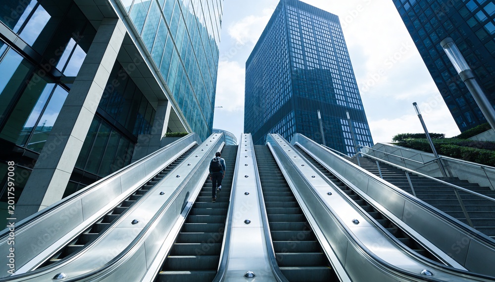 Fototapeta premium Ascending center escalator commuter wearing dark jacket and backpack moving up plaza, 3 steel banks
