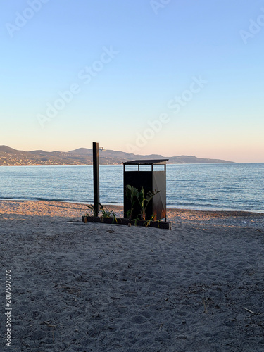 Tranquil beach sunset view with a silhouette of a beach changing room on the sand, calm sea and coastal mountains in the background at twilight.