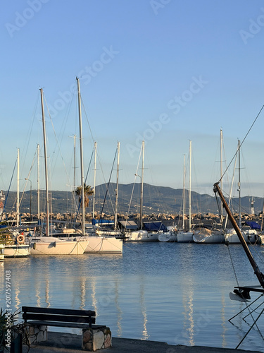 Scenic view of a peaceful marina with white sailboats docked in calm blue water, coastal mountains and clear sky during the golden hour. Greece