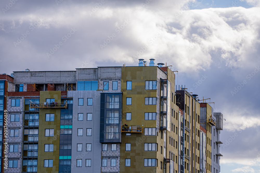 Fototapeta premium High-rise apartment building under construction, featuring multiple facades, thermal insulation, windows, and exterior finishing work in progress beneath a cloudy sky, symbolizing urban development