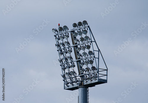 High stadium lighting mast with powerful floodlights against a cloudy sky.
