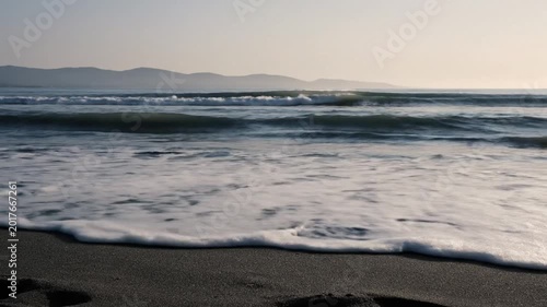 Serene coastal beauty unfolds as foamy waves gently kiss the dark sandy shore under a soft, hazy sky, offering a tranquil escape