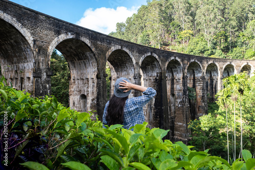 Woman traveler from behind looking at Nine Arch Bridge in Ella, Sri Lanka. Girl holding hat among tea bushes in front of historic stone railway viaduct.