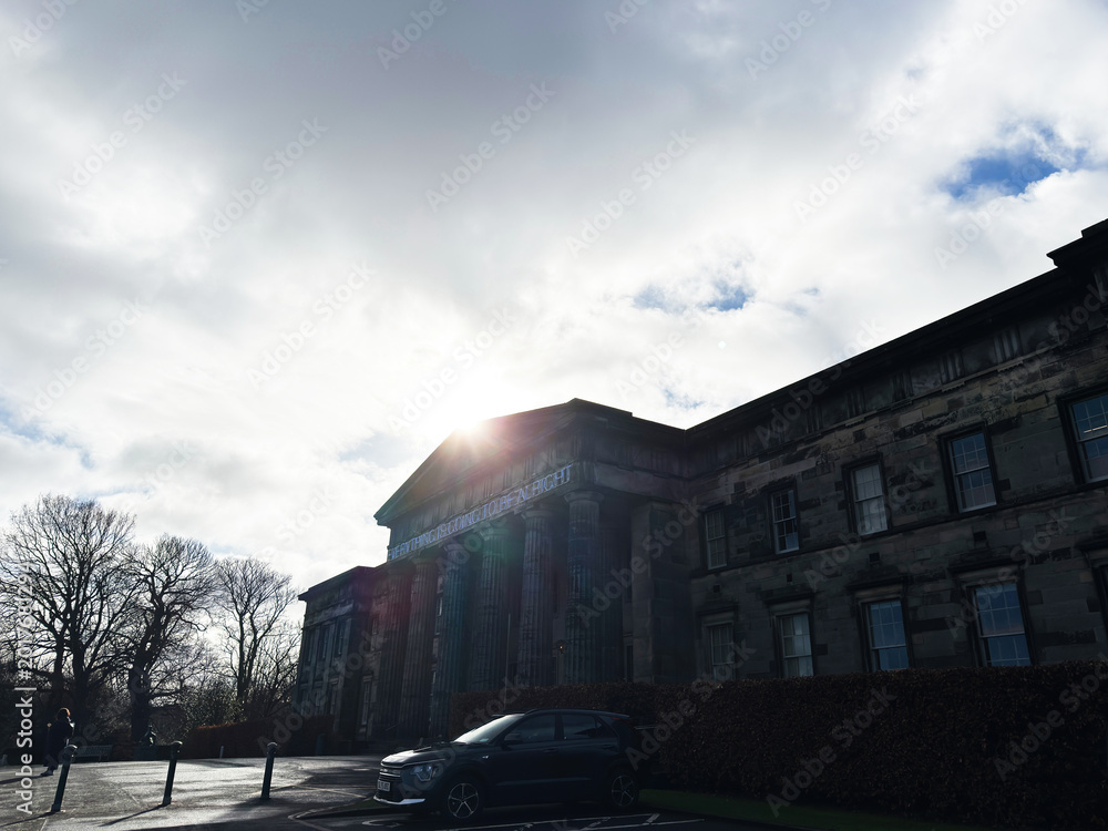 Fototapeta premium Sunlit neoclassical National Galleries Scotland, Modern One building facade with parked car and bare trees in Edinburgh, Scotland, UK under a dramatic cloudy sky