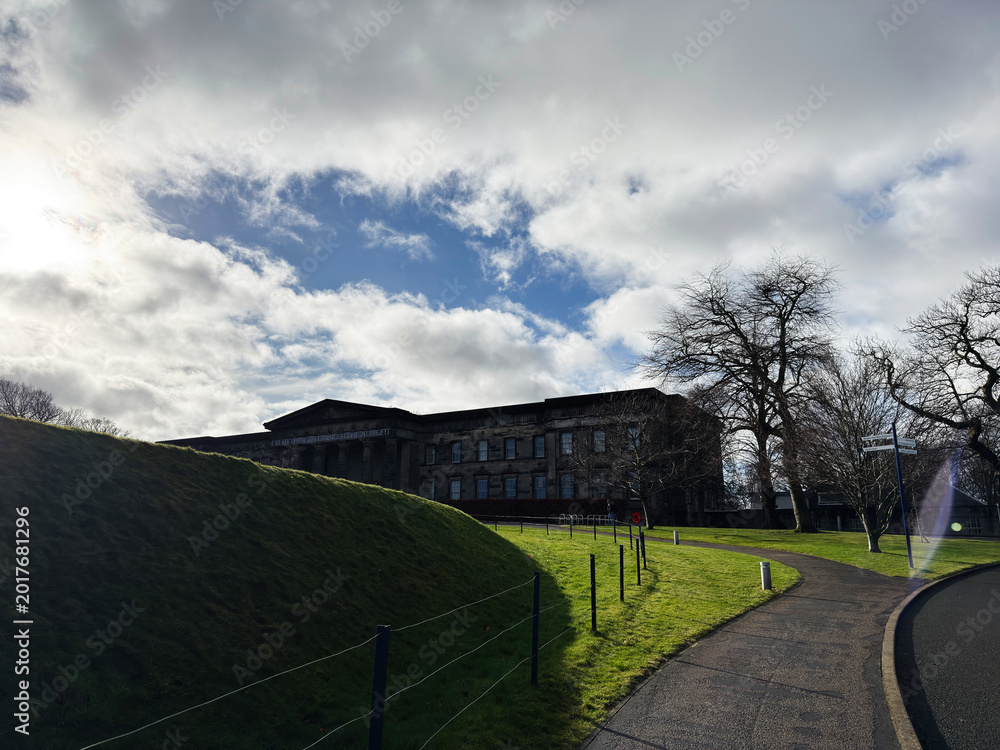 Fototapeta premium Sunlit neoclassical National Galleries Scotland, Modern One building on a grassy hill with winding path and bare trees in Edinburgh, Scotland, UK under a dramatic cloudy sky