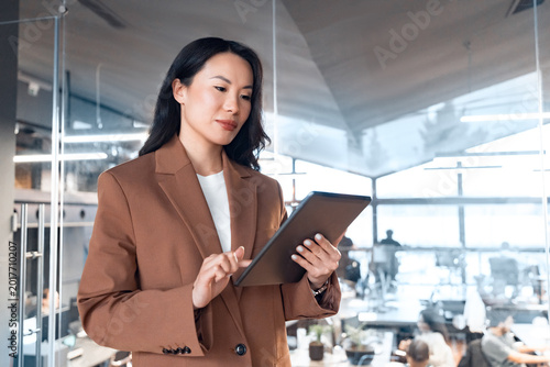 Successful focused Asian mid age woman in suit using pad computer for business app. Busy executive CEO businesswoman professional holding digital tablet pc device for work in modern office workspace