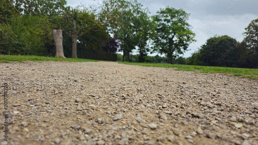 Fototapeta premium Low angle view of a wide gravel pathway leading through a green park with trees under a cloudy sky, England, UK