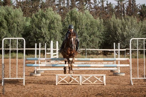 Young equestrian rider jumping over hurdles on a chestnut horse in an outdoor sand arena, surrounded by lush green olive and palm trees. Action shot of show jumping training session on a sunny day.