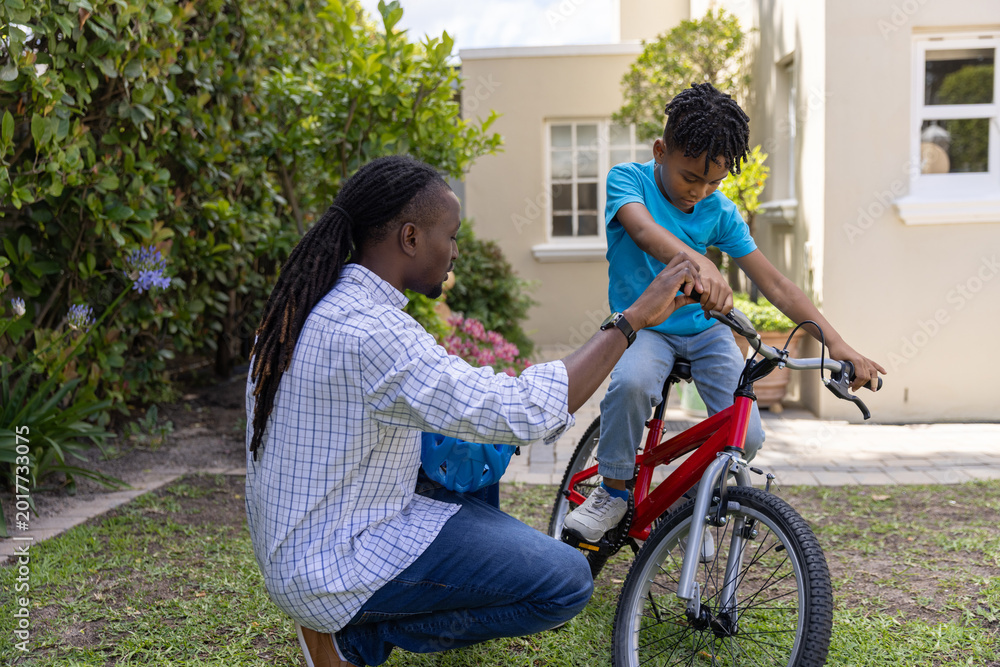Naklejka premium African American man and child boy kneeling front yard steadying red bike, holding blue helmet