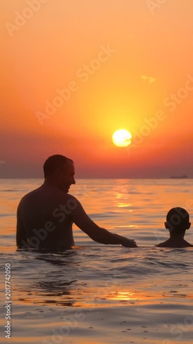 Father and son enjoying sunset in water. Dad and child share joyful moments and laughter at beach. Warm atmosphere creates memorable family bonding experience. Vertical shot captures beautiful scenery