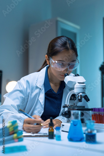 Fotografie Female scientist wearing safety goggles, looking through microscope and writing