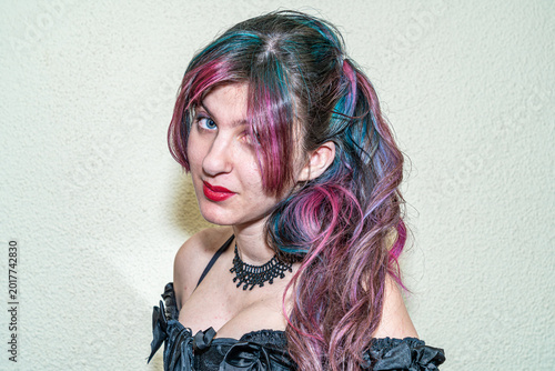 Close-up portrait of a young woman wearing gothic style clothing and red lipstick, showcasing a creative multicolored hairstyle with pink, blue and purple tones against a neutral indoor background.