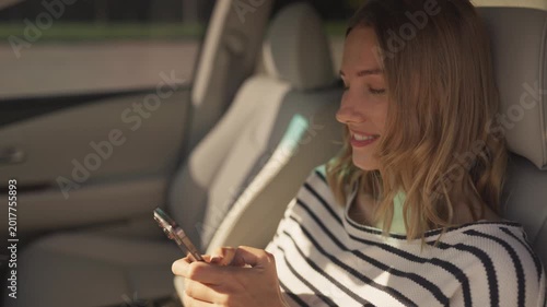 Smiling woman in black-and-white striped sweater sitting in car, holding smartphone and chatting. Looking at screen of mobile phone with happy expression, sunlight illuminating interior. Texting SMS.