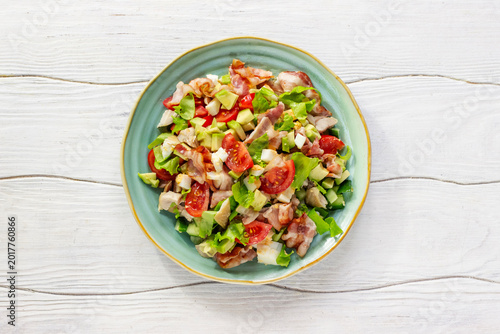 Cobb salad with chicken, avocado, tomatoes, eggs and bacon served on a light blue ceramic plate on a white rustic wooden table.