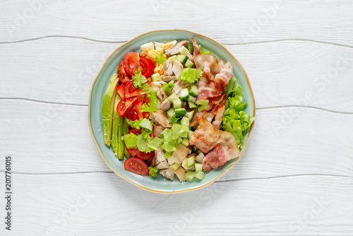 Cobb salad with chicken, avocado, tomatoes, eggs and bacon served on a light blue ceramic plate on a white rustic wooden tabletop.