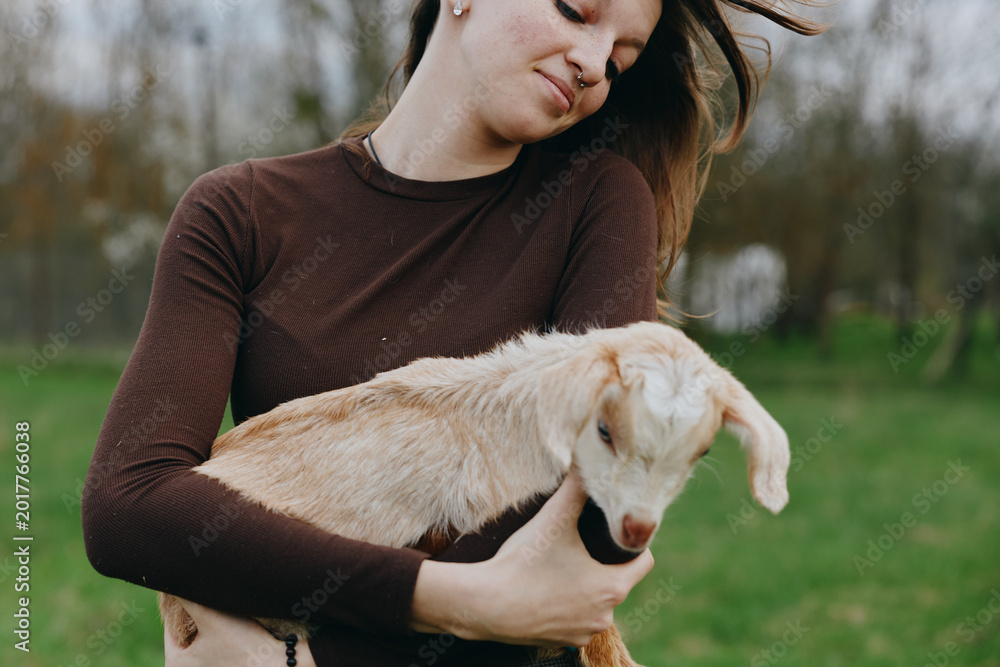 Naklejka premium Lifestyle moment with a woman outdoors, gently holding a goat kid, warm sunlight, casual sweater, soft breeze, grassy field, natural colors, peaceful bond and connection