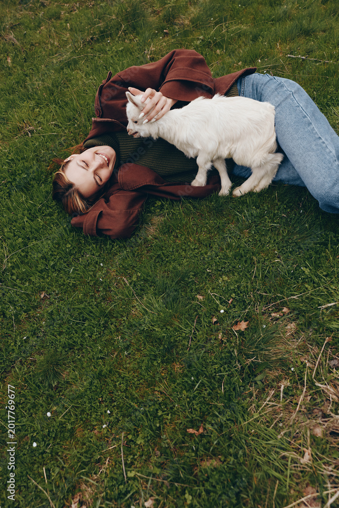 Naklejka premium Cheerful woman outdoors, lying on grass, goat kid nuzzles her hand, relaxed pose, sunlit scene, natural colors, gentle breeze, countryside mood, authentic lifestyle moment, joyful connection