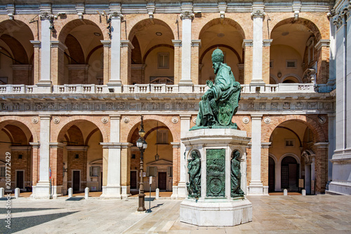 Bronze statue of Pope Sixtus V sitting on a marble pedestal, adorned with reliefs, standing in front of the ornate Palazzo Apostolico with its arches in Loreto, Marches, Italy