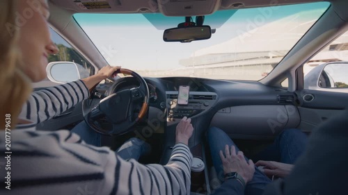Caucasian woman driving car using GPS navigation on smartphone while passenger sits beside. Female driver holding steering wheel and adjusting map in daylight. Rear. Learning to drive. Back view.