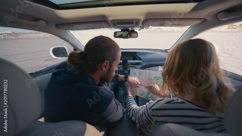 Man with tied hair driving car while female passenger holds paper map and points at location. Couple navigating route together inside vehicle during road trip in daylight through open parking area.