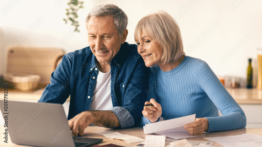 © Prostock-studio - A couple is sitting at a kitchen table looking at a laptop. They appear to be discussing financial matters while holding papers, looking engaged and focused on their task.