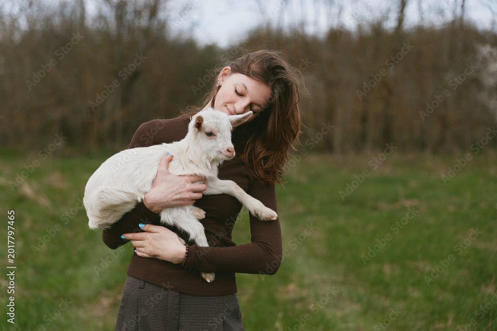 Naklejka premium A woman in a brown sweater cradles a white goat kid in a sunlit meadow, smiling softly as they share a quiet moment. A lifestyle scene of animal bonding and care.