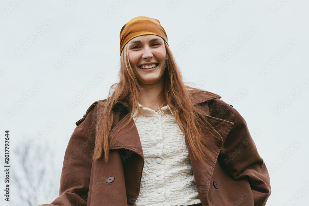 Obraz premium Cheerful woman outdoors, smiling, wearing a warm brown coat and bandana, casual pose, natural colors, soft breeze, sunny atmosphere, candid lifestyle portrait, autumn tones, relaxed vibe, simple