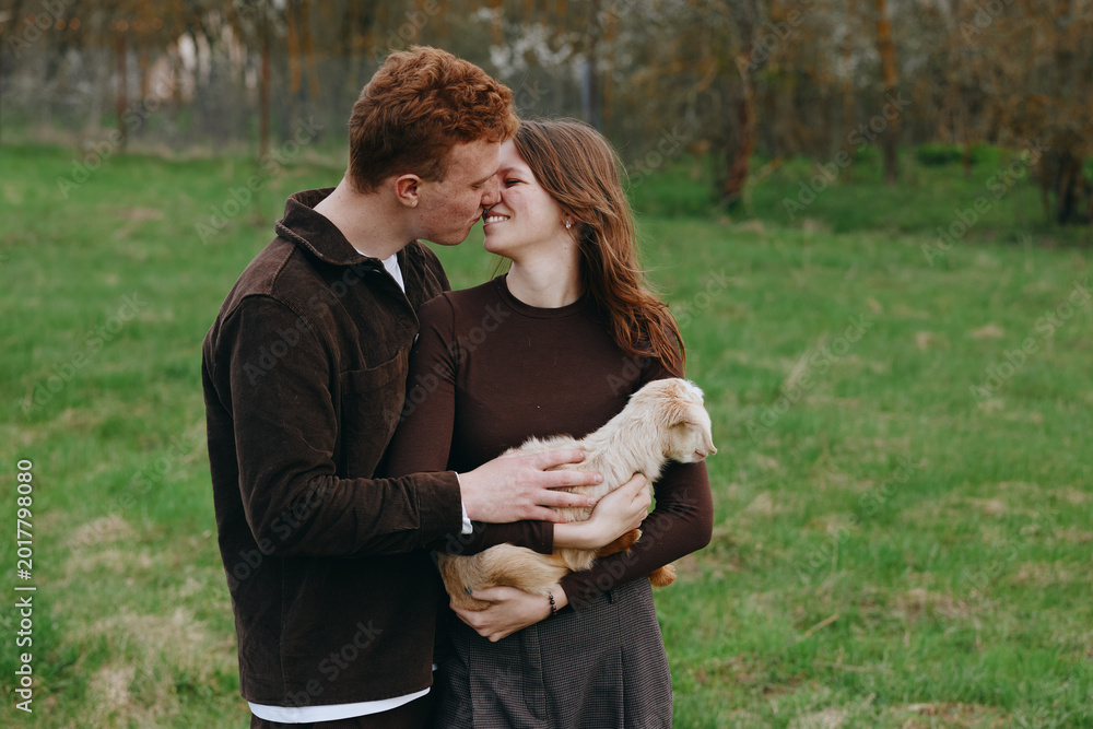 Naklejka premium Happy couple in a countryside field cuddle a young goat kid, sharing tender smiles and hands on the small animal, capturing warm lifestyle moment with gentle farm charm.