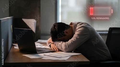An exhausted male employee sleeping on an office desk with a laptop and documents showing a low battery