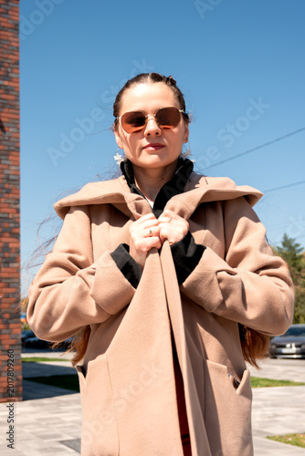 Stylish woman in camel coat standing outdoors against blue sky and brick wall. Street fashion portrait concept for elegance, warmth and urban city lifestyle.
