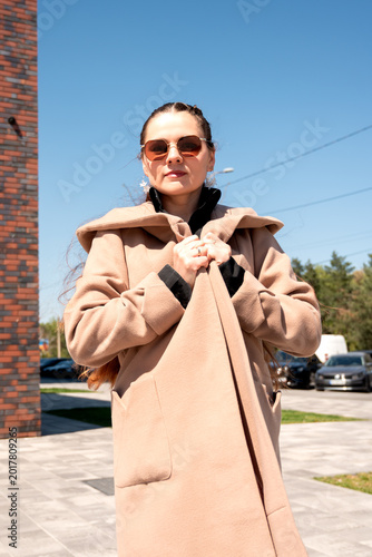 Confident young woman in oversized beige coat holding collar on sunny city street. Urban autumn fashion concept for elegance and modern female lifestyle.