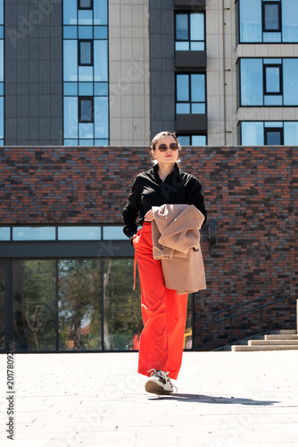 Confident woman in bold red pants holding coat in sunny urban setting. Street style fashion concept for female independence and modern city lifestyle.