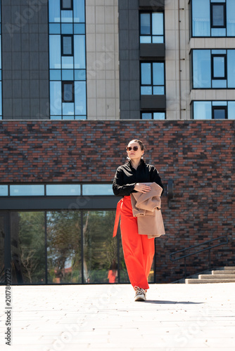 Stylish young woman in red wide-leg trousers carrying beige coat over arm in front of modern glass building. Urban street fashion and confidence concept.
