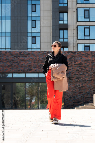 Urban fashion portrait of a woman in striking red and black outfit holding beige coat against modern architecture. Concept of style, freedom and independence.