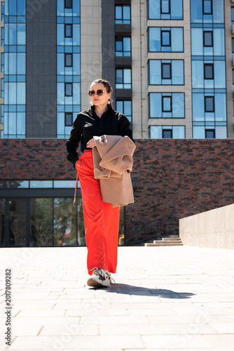 Determined young woman in vibrant red wide-leg pants with coat in hand standing in urban plaza. Bold street style concept for female confidence and city life.