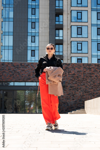 Poised young woman in red palazzo trousers carrying coat in sunny city square. Contemporary street style concept for modern urban woman and fashion lifestyle.