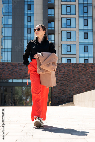 Full length portrait of a stylish woman in red trousers and black shirt carrying beige coat outdoors. Modern city fashion concept for confidence and elegance.