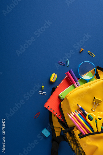 Back to school vertical essentials: yellow backpack with colorful stationery and notebooks on blue background