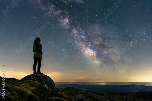 Silhouette of a person on a rocky hill under a starry Milky Way sky at twilight over a vast landscape