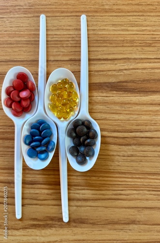Vertical top view image of colorful capsules and supplements arranged in ceramic spoons over a wooden table. Concept of healthcare, nutrition, healthy lifestyle, vitamins, pharmaceutical products