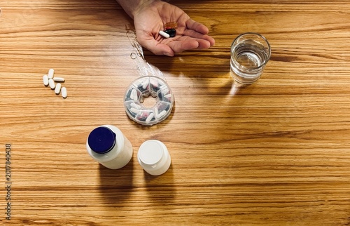 Top view of assorted medicines, capsules, and tablets arranged near medicine bottles and a glass of water while a person takes medication by hand. Healthcare, pharmaceutical therapy, and medical treat