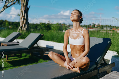 Photography A woman sits cross legged on a poolside lounge, eyes closed, basking in sunlit calm