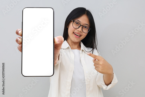 A cheerful young Asian woman wearing casual clothes, holding a mobile phone, pointing at a blank screen of the phone, smiling while looking at the camera, standing isolated on a white background.