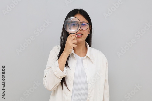 A young Asian woman wearing casual clothes, holding a magnifying glass is looking for something with a serious expression, isolated on a white background.