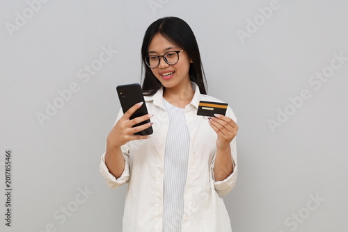 A cheerful young Asian girl smiles, shows a credit card and a smartphone, recommends banking services via mobile, standing on a white background.