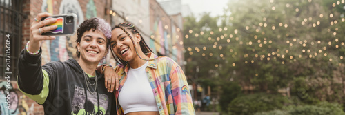 Young LGBT couple taking a selfie on the street. Diverse lesbian women smiling with rainbow pride flag on phone. Wide banner with copy space for text